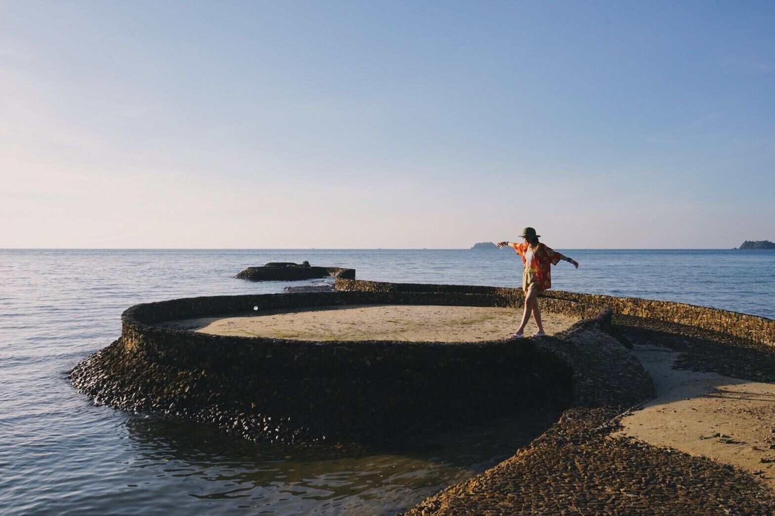 A person standing on a circular stone platform by the sea with a clear sky.