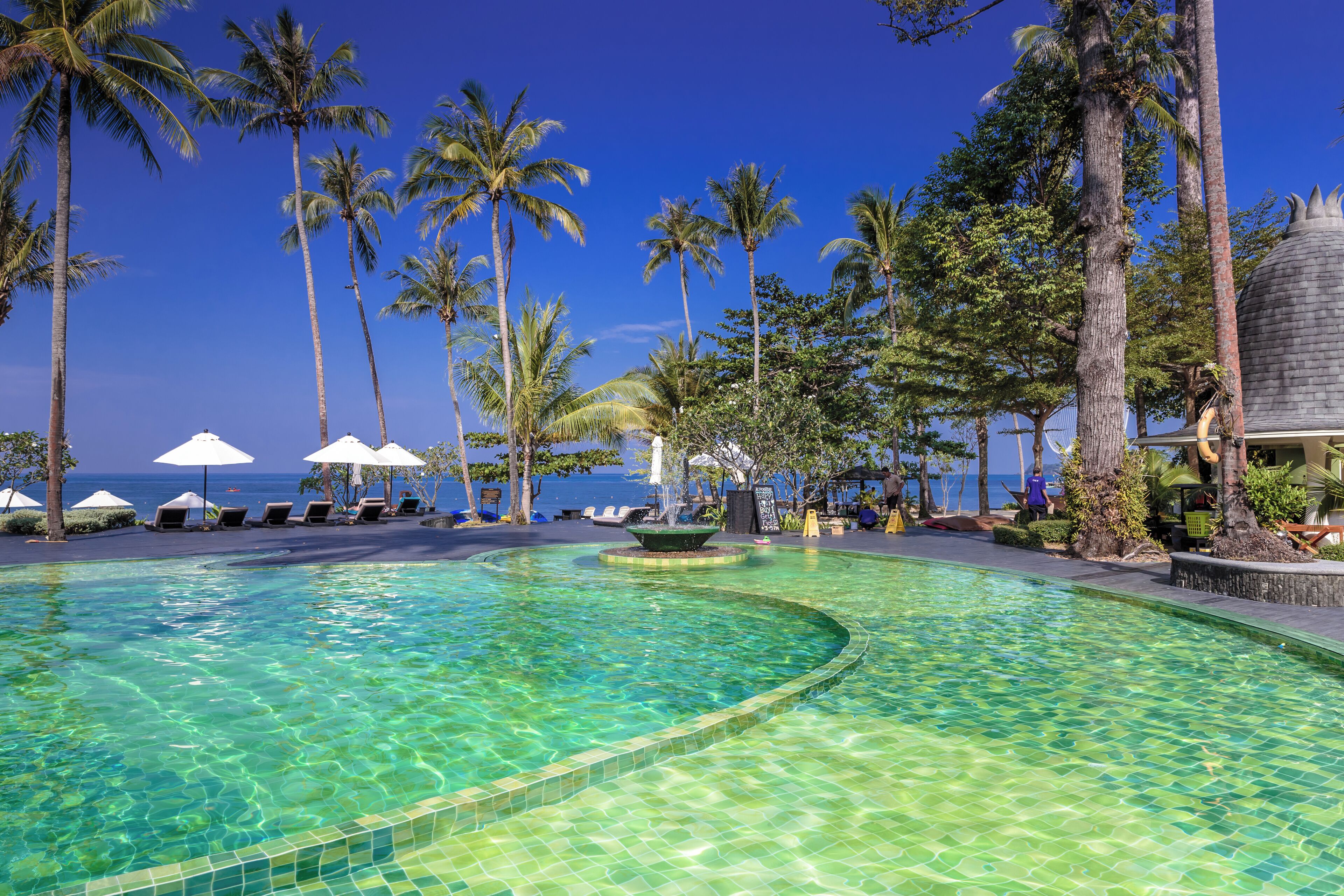 Una piscina cristalina con bordes de azulejos, rodeada de sillones de descanso y sombrillas, con palmeras y vistas al mar.
