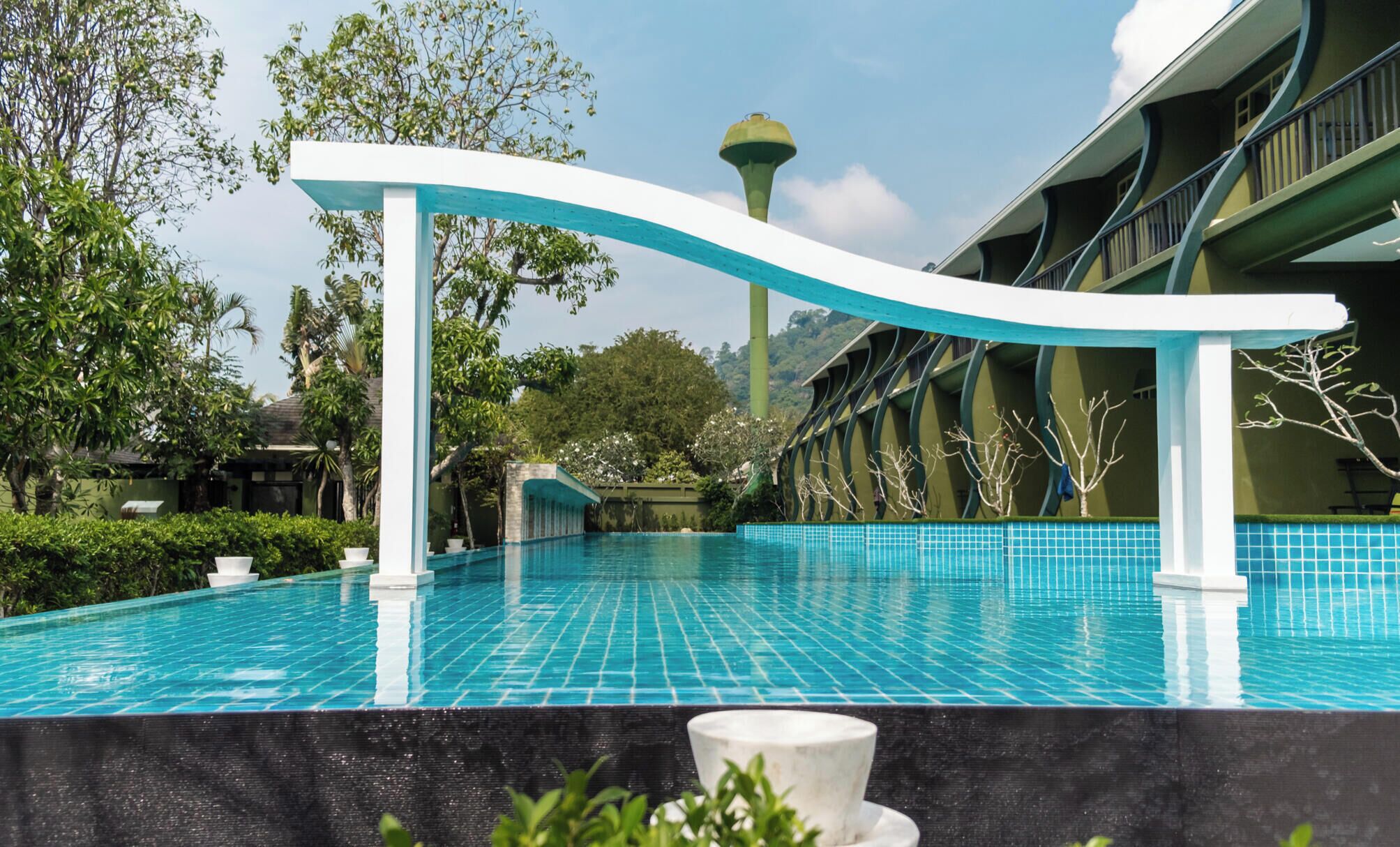 A swimming pool with a curved white structure, surrounded by greenery and a building with balconies.