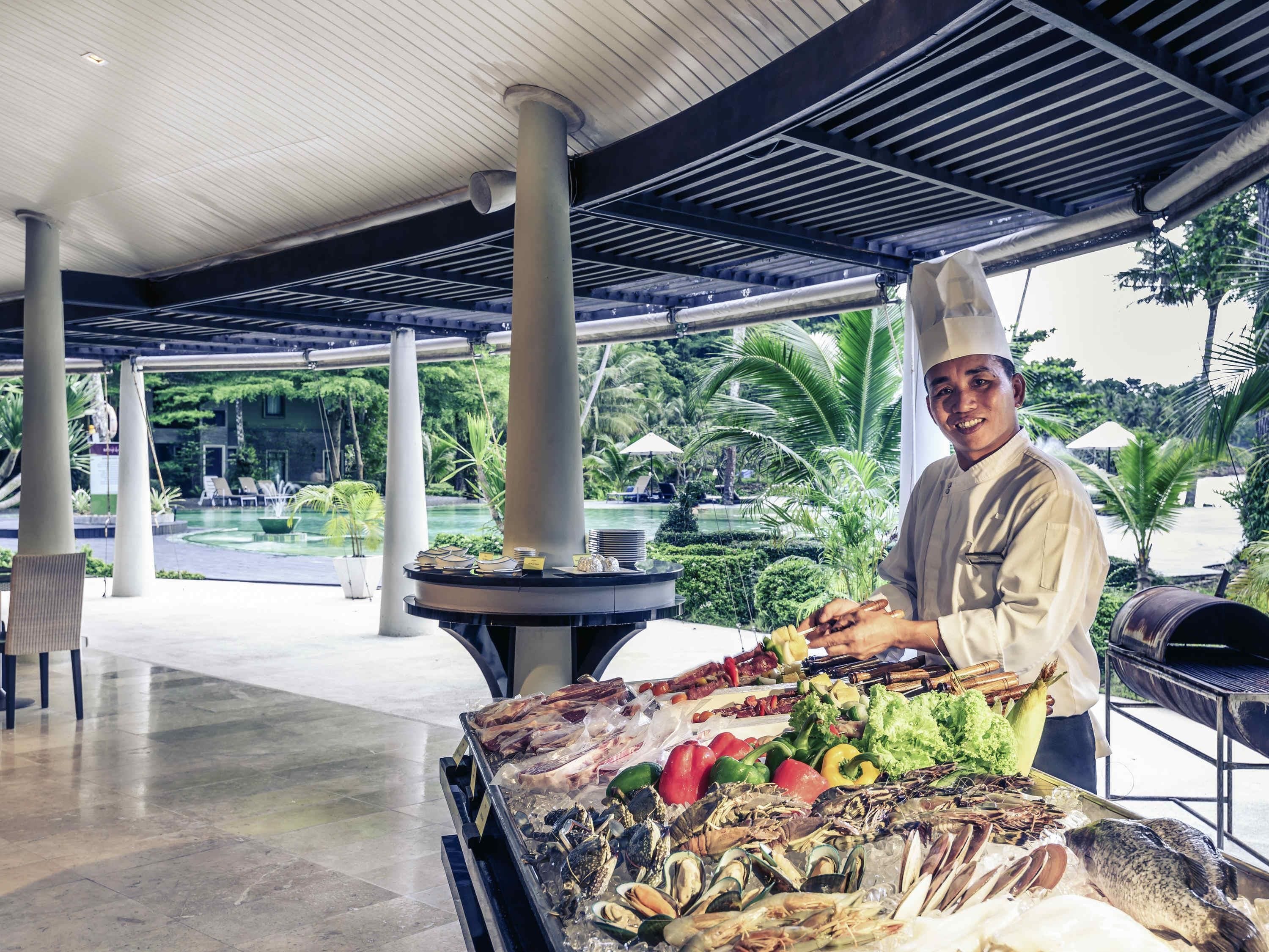 Un chef preparando mariscos en un área de comedor al aire libre cubierta, con una piscina y follaje al fondo.