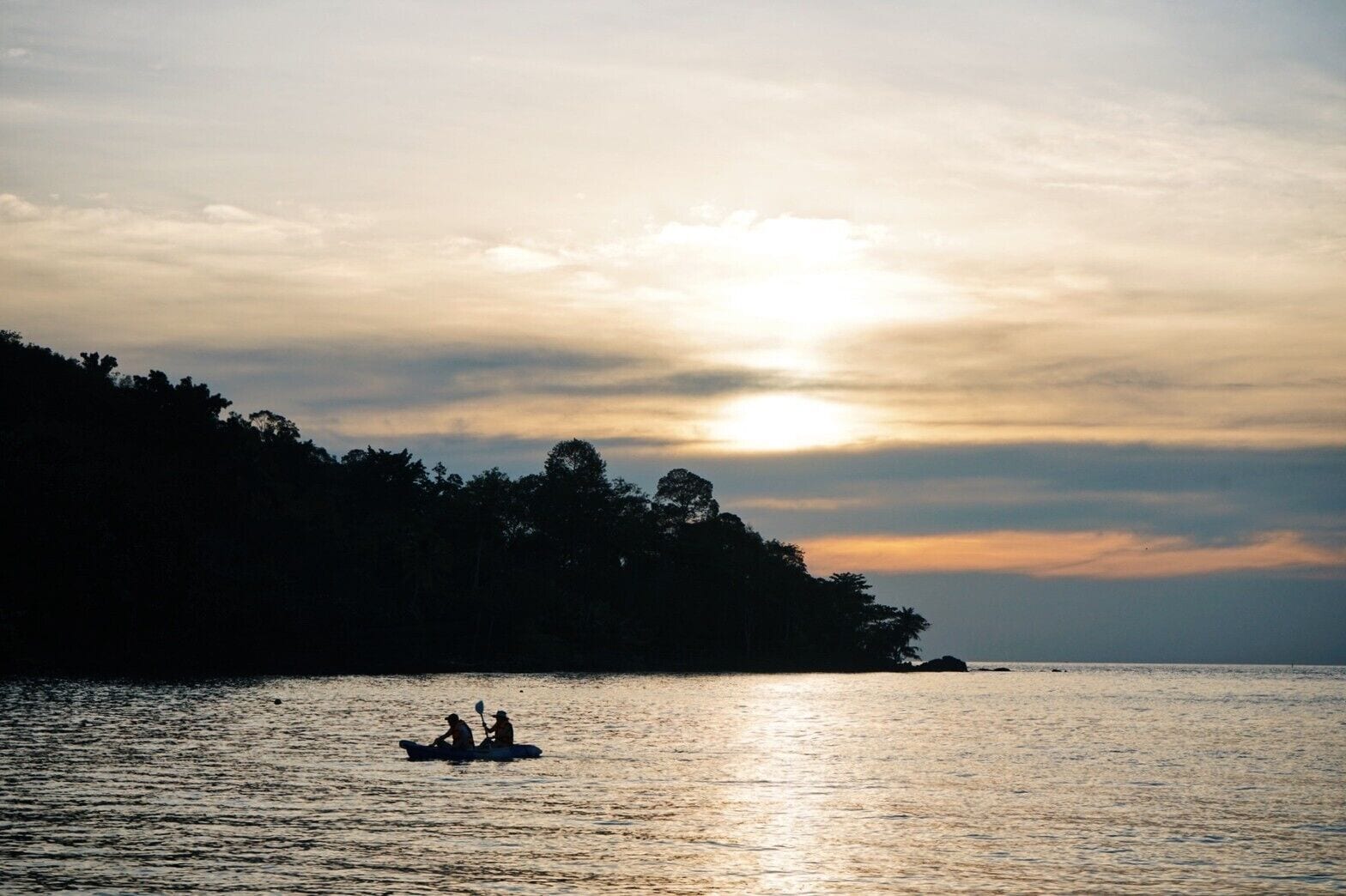 Un plan d’eau calme, avec un bateau transportant deux personnes, entouré d’une côte boisée et avec un coucher de soleil en arrière-plan.