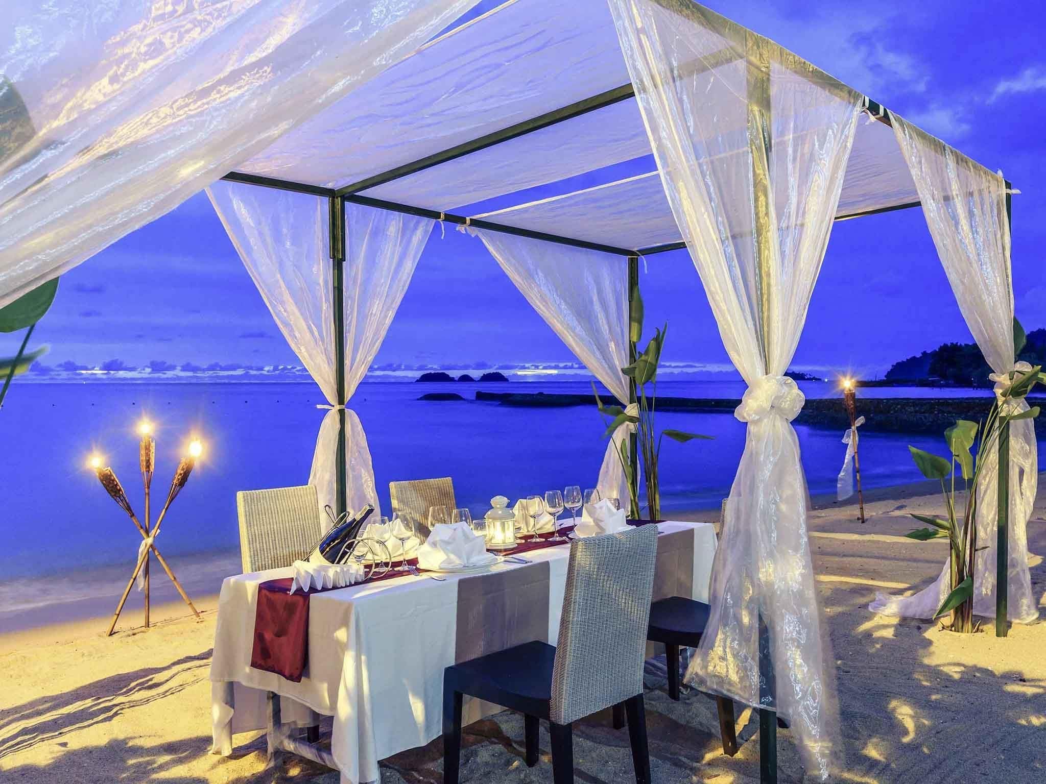 A beachside dining setup with a canopy, table, chairs, and decorative lighting.