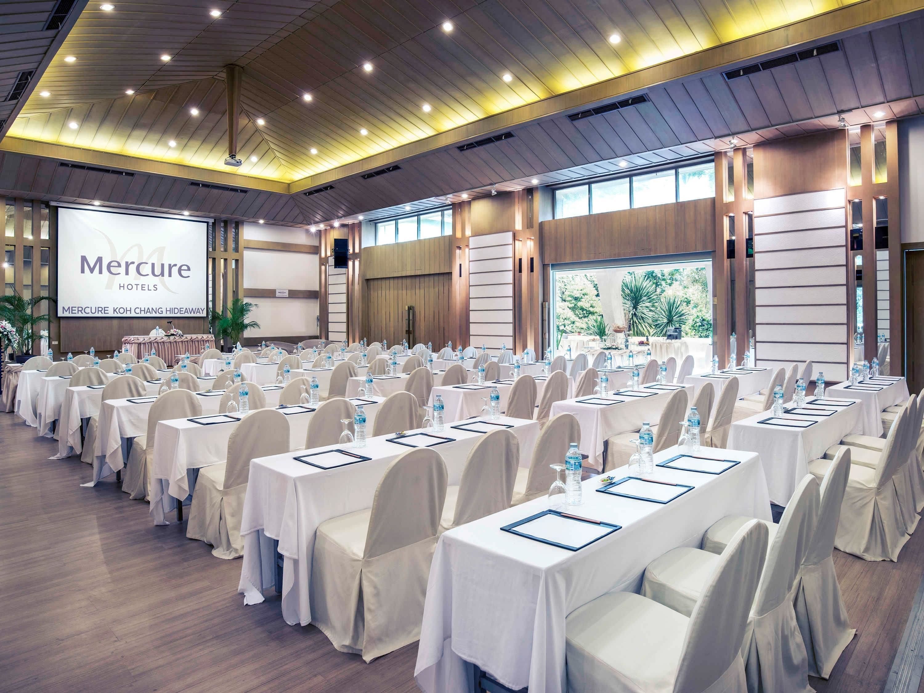 Un salón de conferencias en Mercure Koh Chang Hideaway con filas de mesas y sillas, una pantalla grande y vistas a la vegetación exterior.