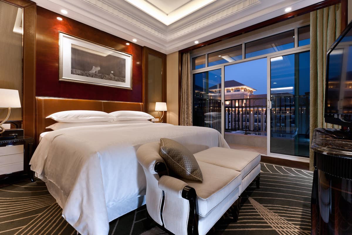 Habitación de hotel con una cama grande, un sofá, un escritorio con lámpara y vista a un edificio a través de la ventana.