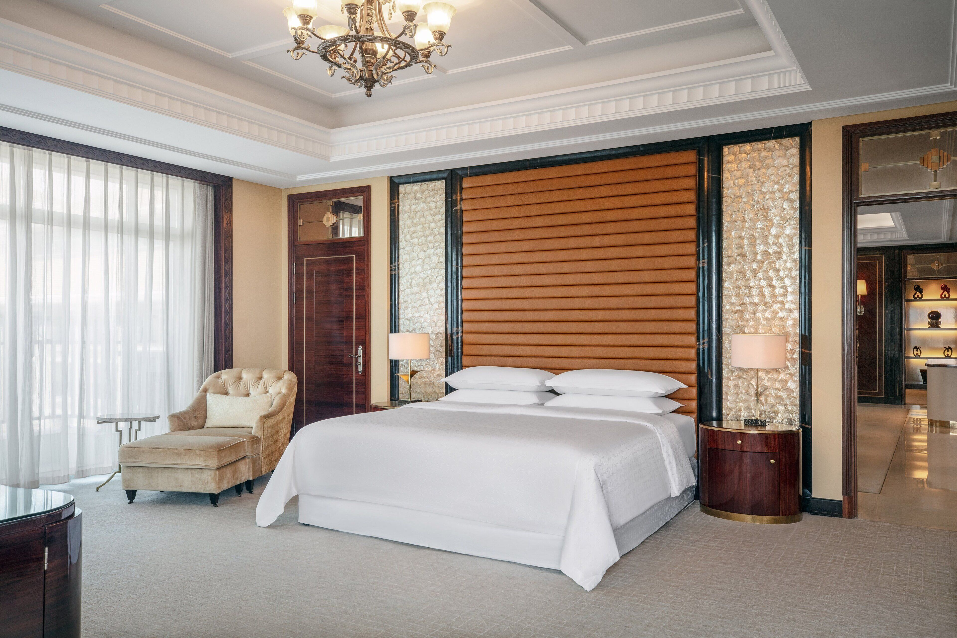 A spacious hotel room with a large bed, a sitting armchair, a small table, and a chandelier.