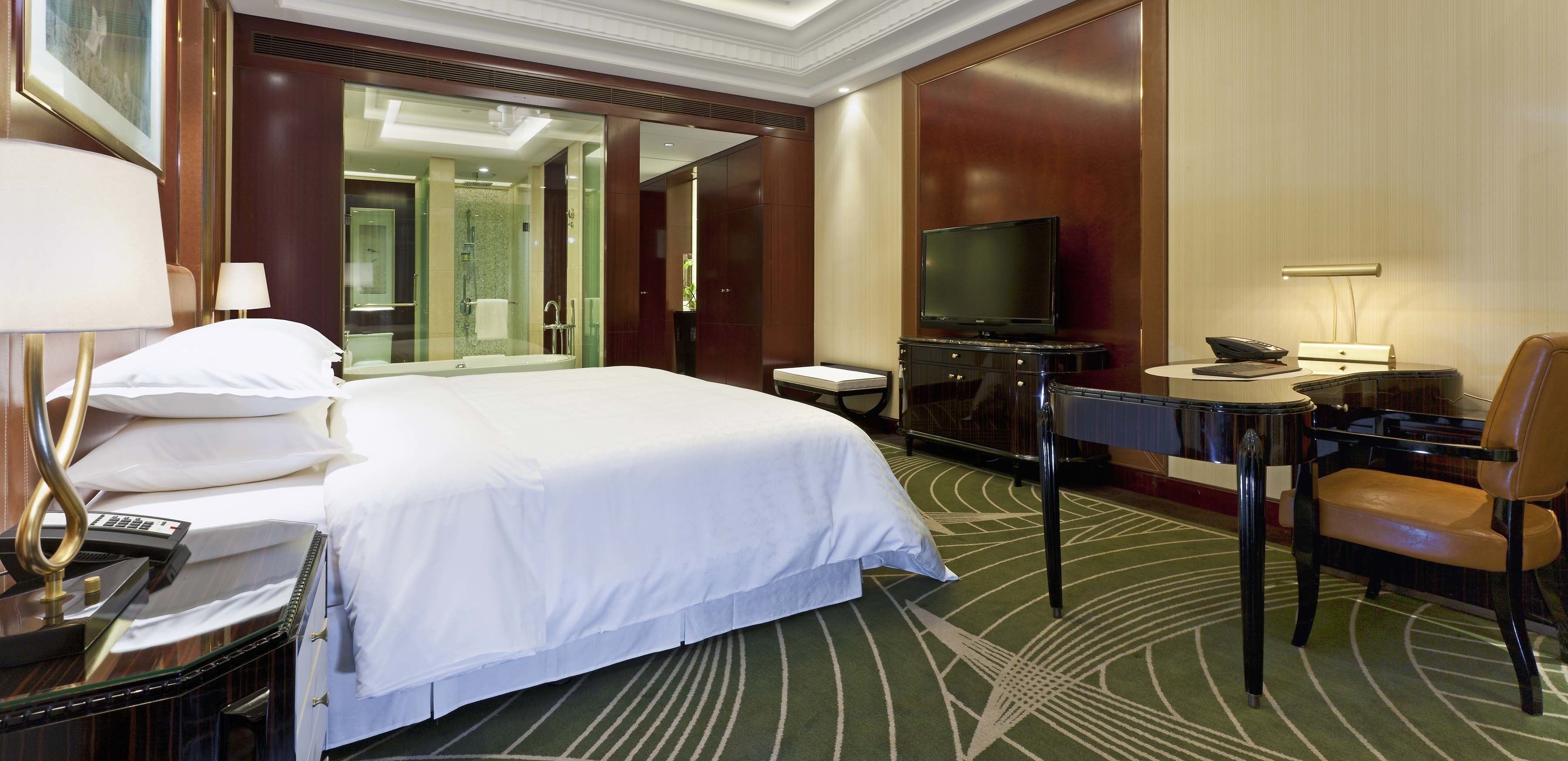 A hotel room with a large bed, a television, a desk with a telephone, and a chair.