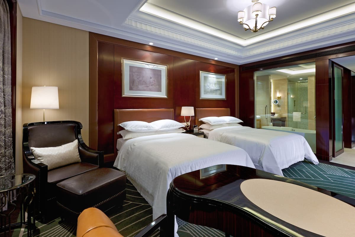 Habitación de hotel con dos camas, una zona de estar con una silla, una mesa y un espejo.