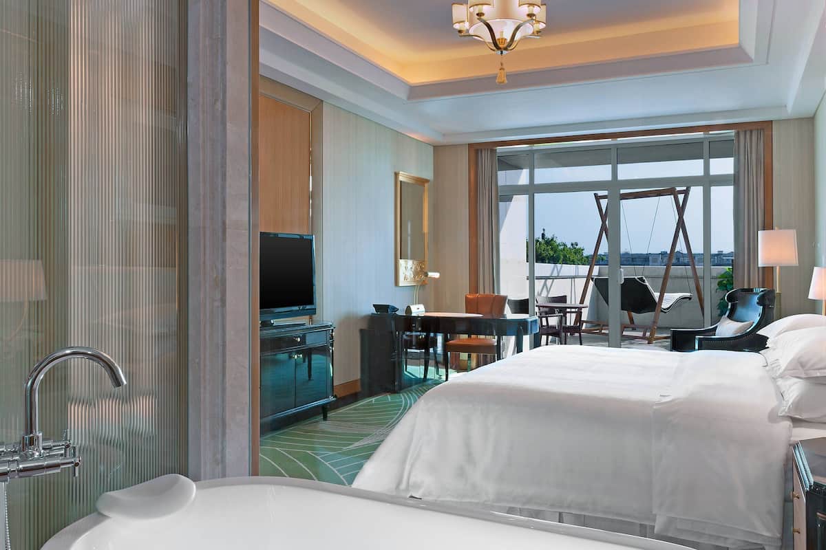 Habitación de hotel con una cama grande, baño con bañera, escritorio con silla, televisor y una ventana grande con vistas.