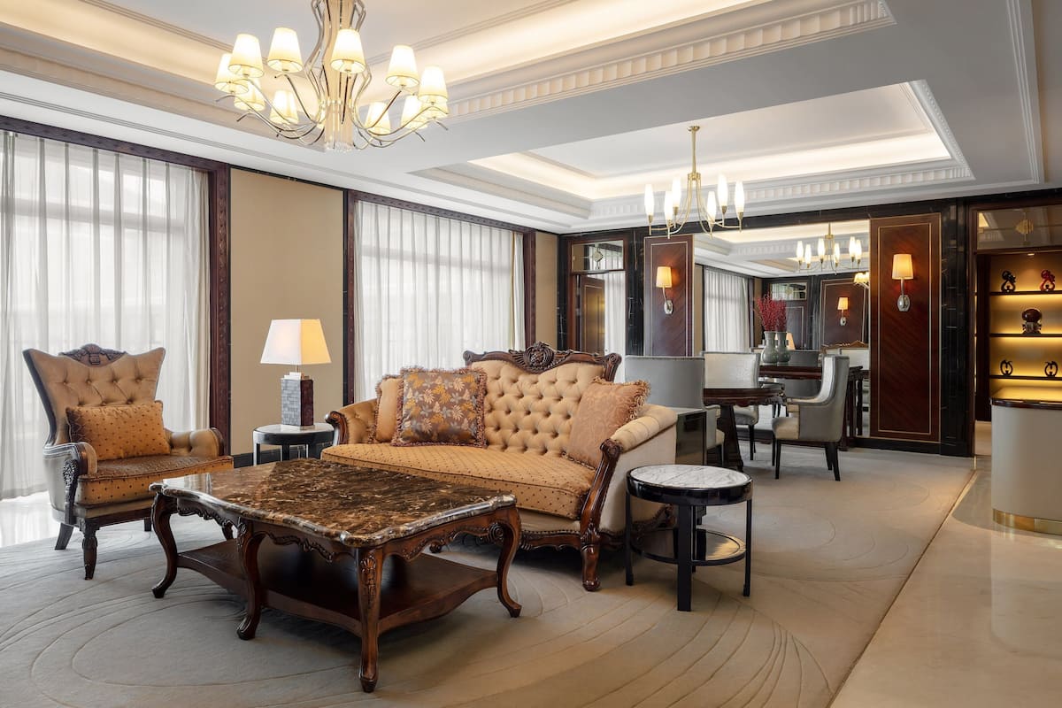 Una lujosa habitación de hotel con una cama grande, dos sillones, una mesa de centro, una mesa de comedor y una araña.