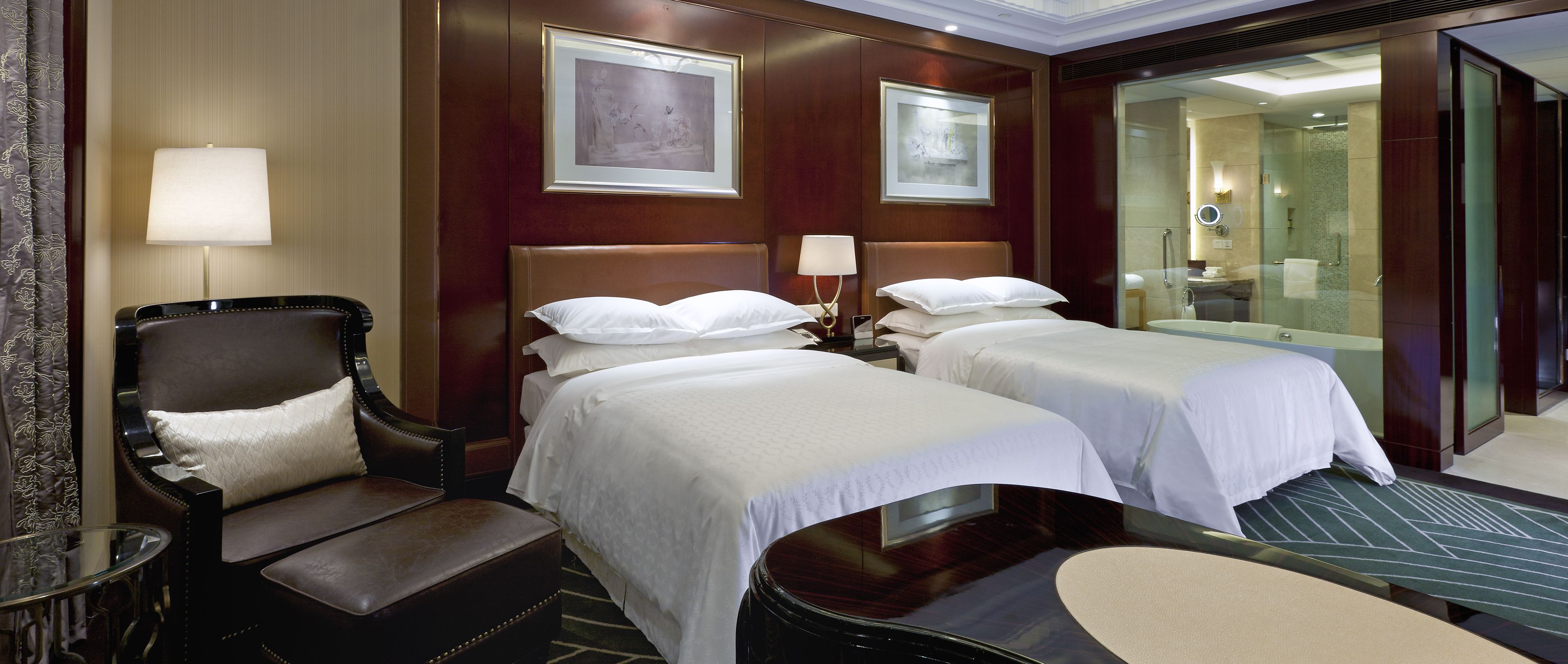 A hotel room with two beds, a sitting area with a chair, a glass-enclosed shower, and framed artwork on the walls.