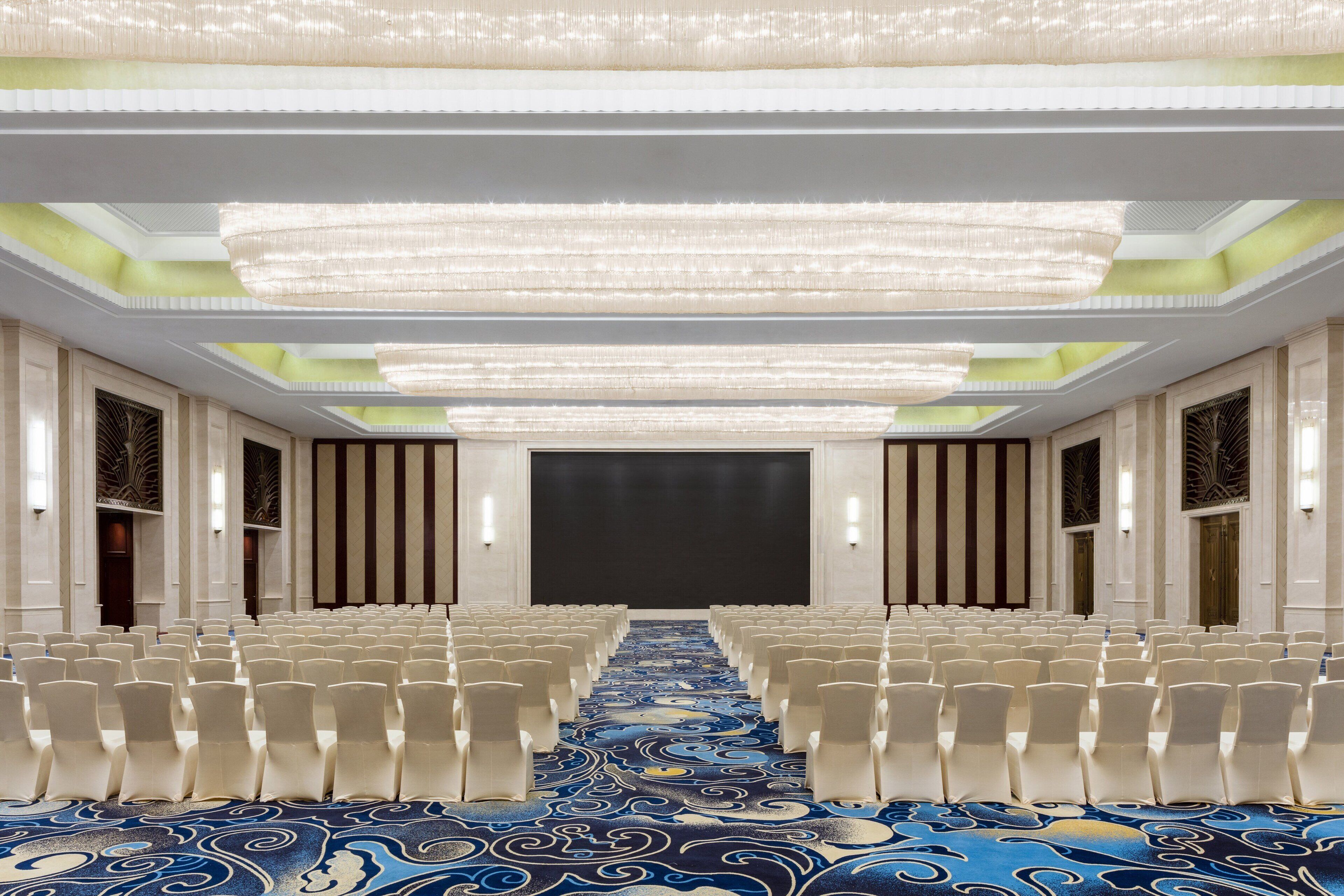 A conference room with rows of white chairs, a patterned carpet, and a large screen at the front.