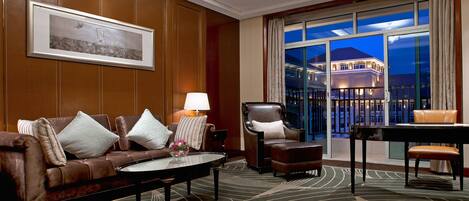 A hotel room with a leather sofa, a coffee table, a chair, and a view of a building through large windows.