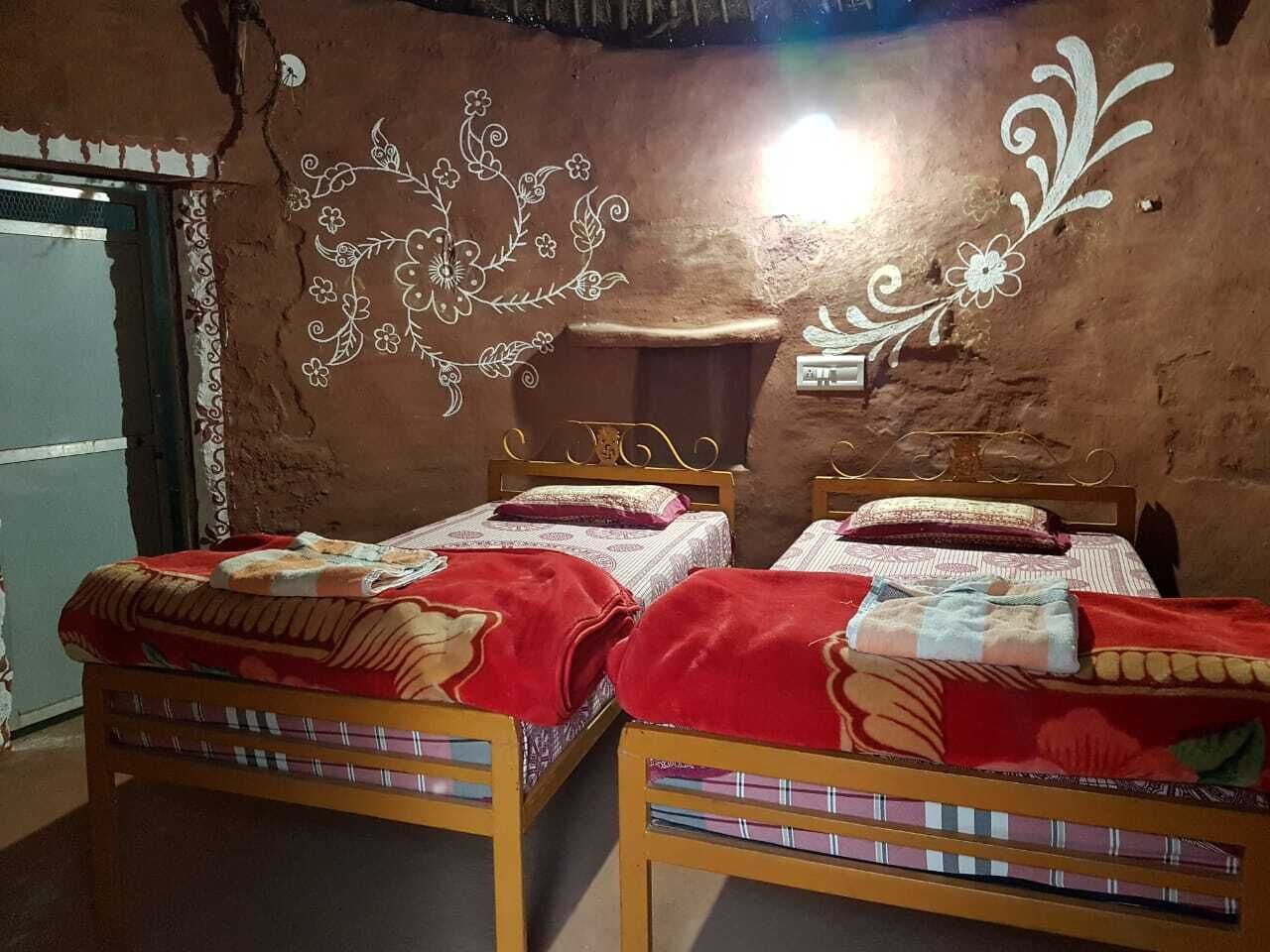 A room with two wooden beds, each with a red blanket and a patterned bedspread. There is a decorative wall with floral designs and a small shelf.