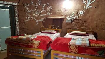 A room with two wooden beds, each with a red blanket and a patterned bedspread. There is a decorative wall with floral designs and a small shelf.