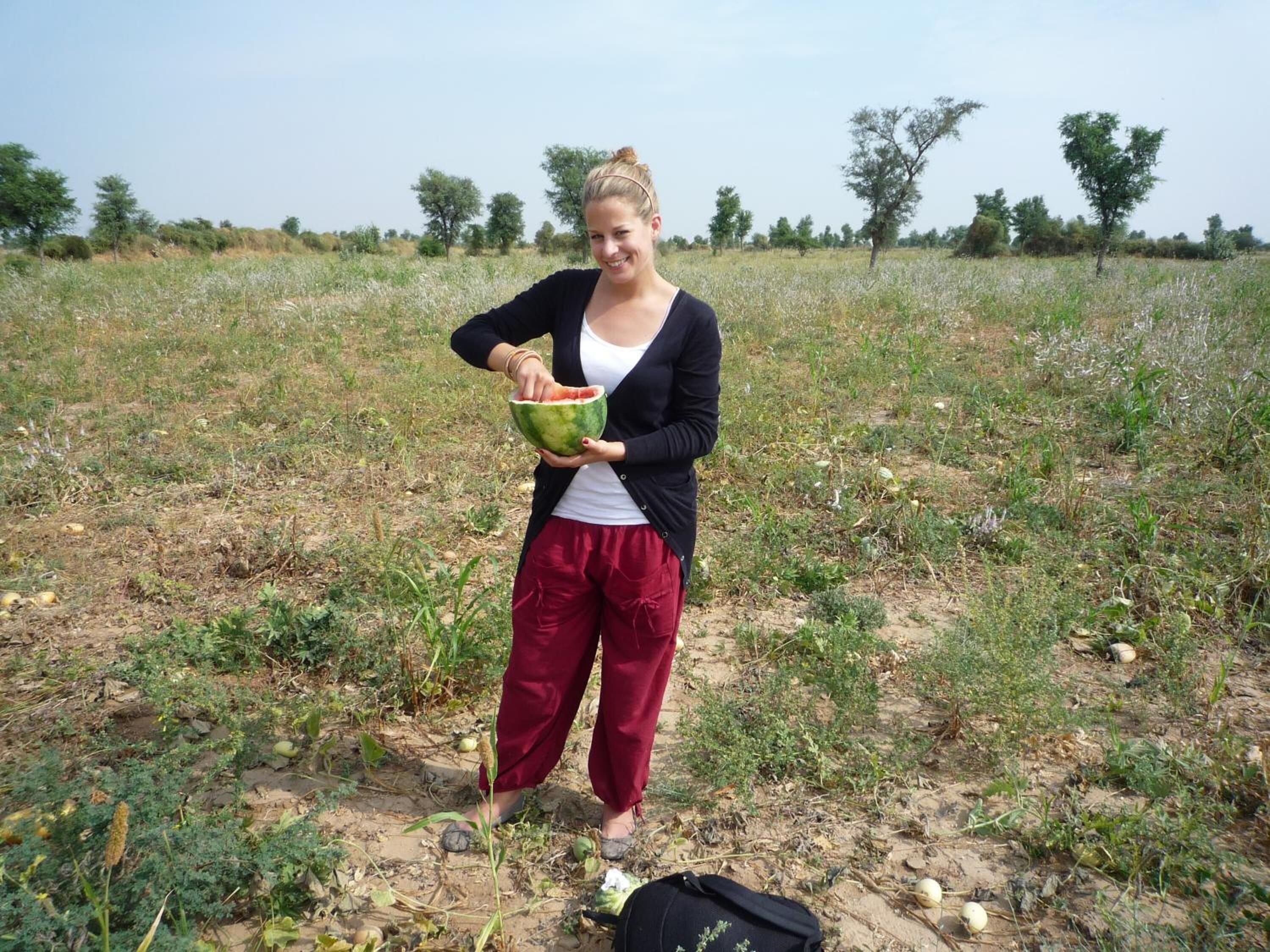 Eine Person hält eine Wassermelone in einer offenen Landschaft mit vereinzelter Vegetation und Bäumen im Hintergrund.
