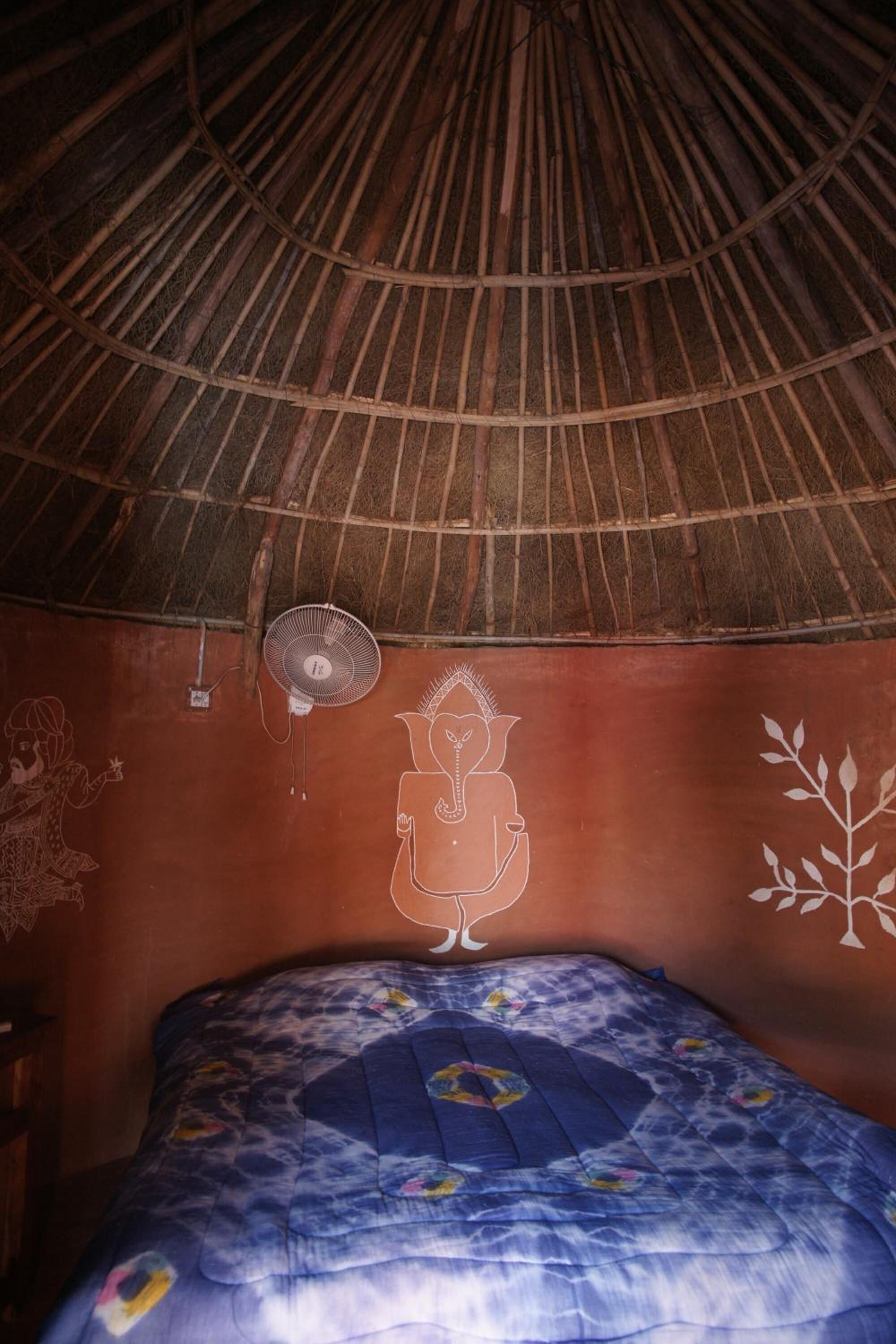 A room with a thatched roof, a bed with a blue floral bedspread, and a wall with a white mural.