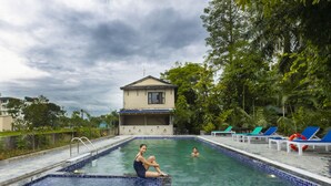 Ein Schwimmbecken mit einer gefliesten Kante, ein Gebäude im Hintergrund und eine Reihe von Liegestühlen am Pool.