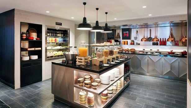 A modern cafe with a central counter displaying various food items and beverages, shelves with condiments and jars, and a clean, tiled floor.
