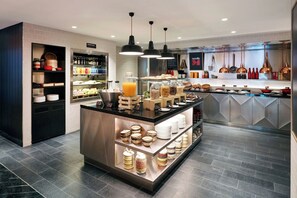 Un café moderne avec un comptoir central où sont exposés divers aliments et boissons, des étagères remplies de condiments et de pots, et un sol carrelé et impeccable.