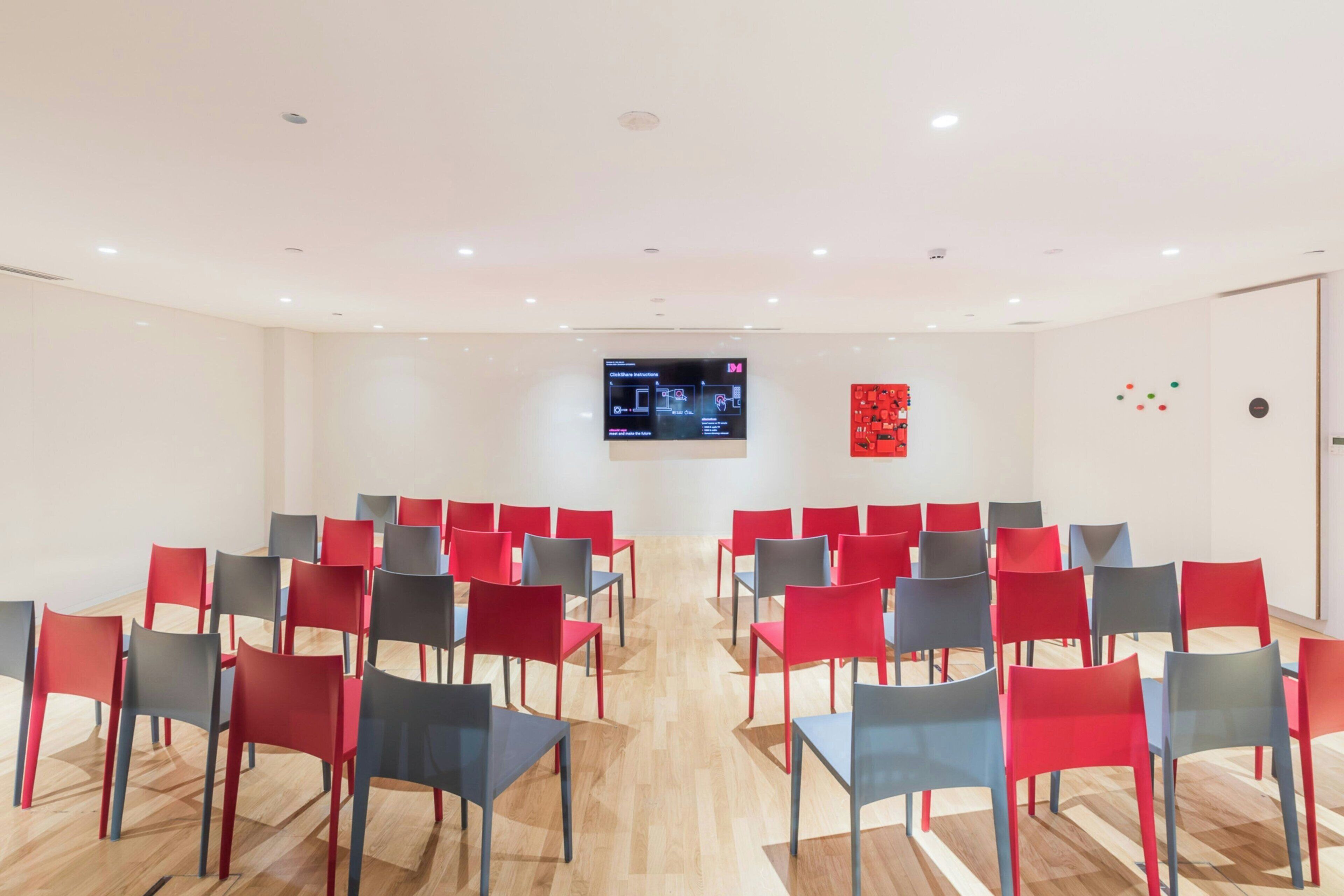 A conference room with rows of red and grey chairs, a large screen at the front, and a red abstract art piece on the wall.