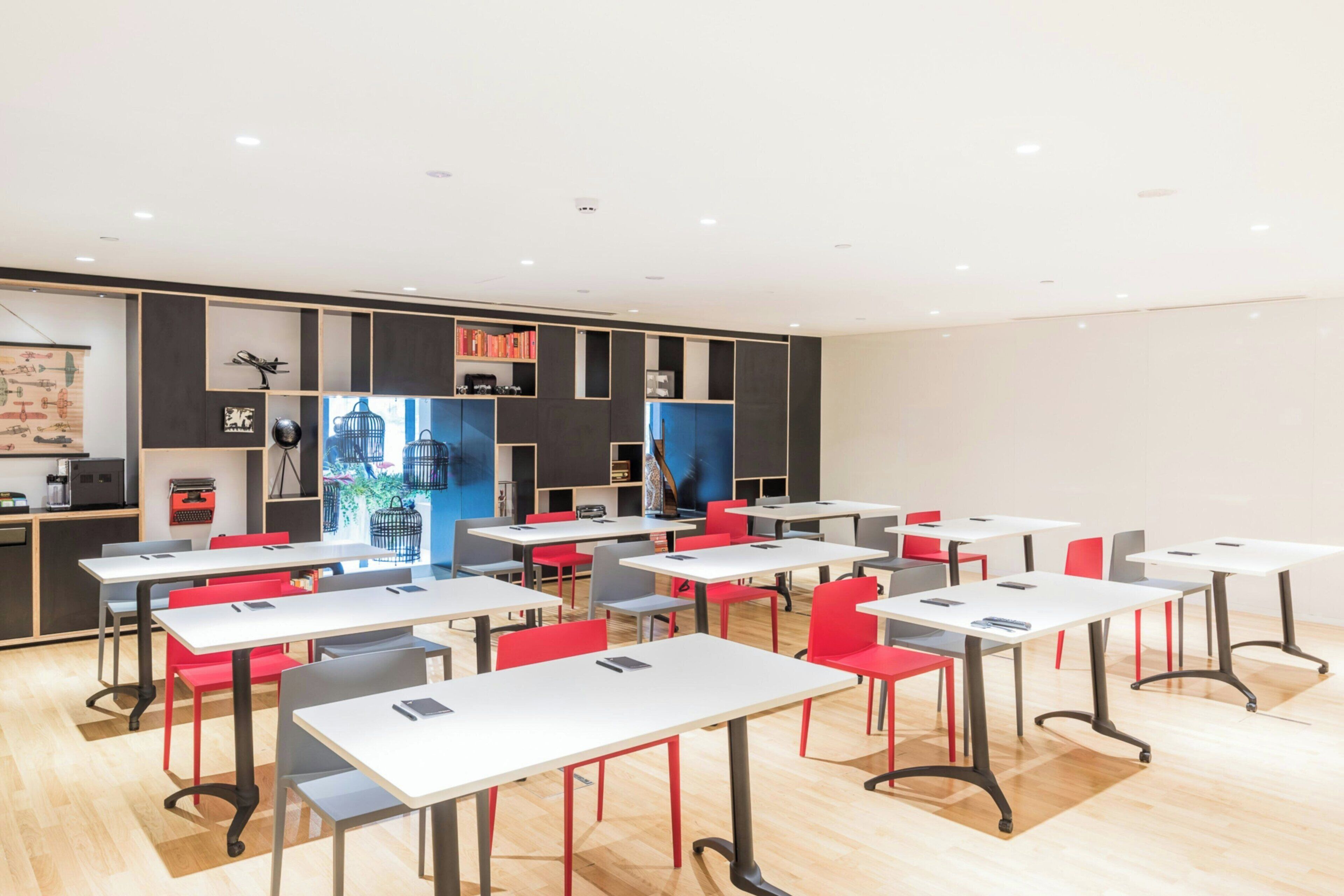 A modern classroom with white tables and red chairs, equipped with laptops and a large window with a view of birds.