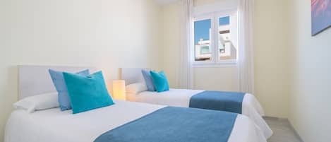 A hotel room with two beds, each with white bedding and blue accents, a window with white curtains, and a view of buildings outside.