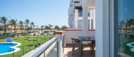 Un balcón con mesa y sillas, con vista a una piscina y palmeras.