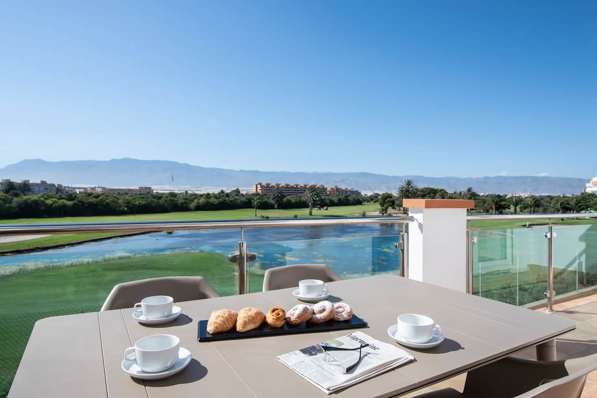 A dining table with croissants and coffee cups, overlooking a pool and mountains.