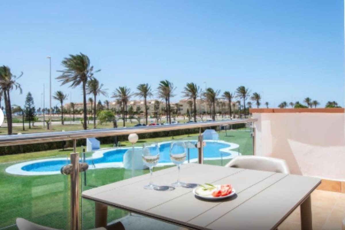 A dining area with a table, two wine glasses, and a plate of food. In the background, there is a swimming pool with palm trees and a clear sky.