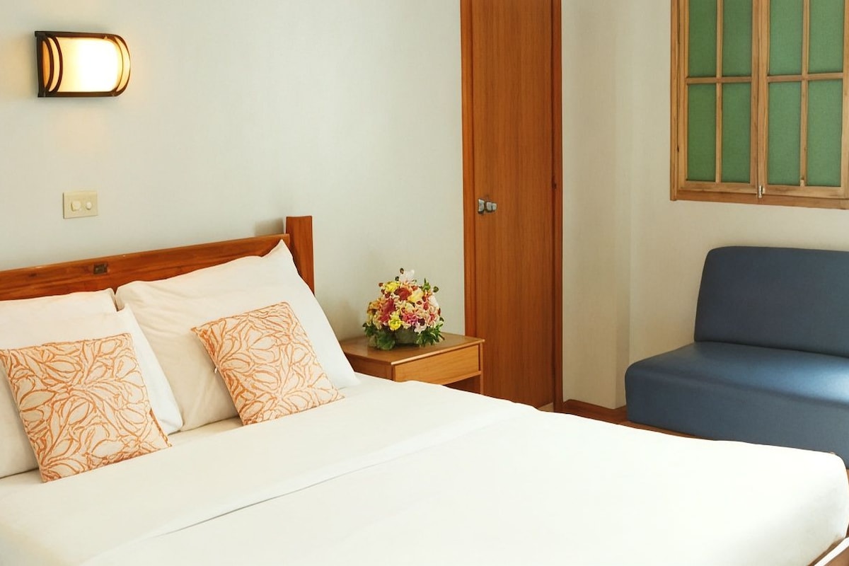 Een hotelkamer met een bed, een nachtkastje met een bloevas, een houten deur en een raam met groene luiken.