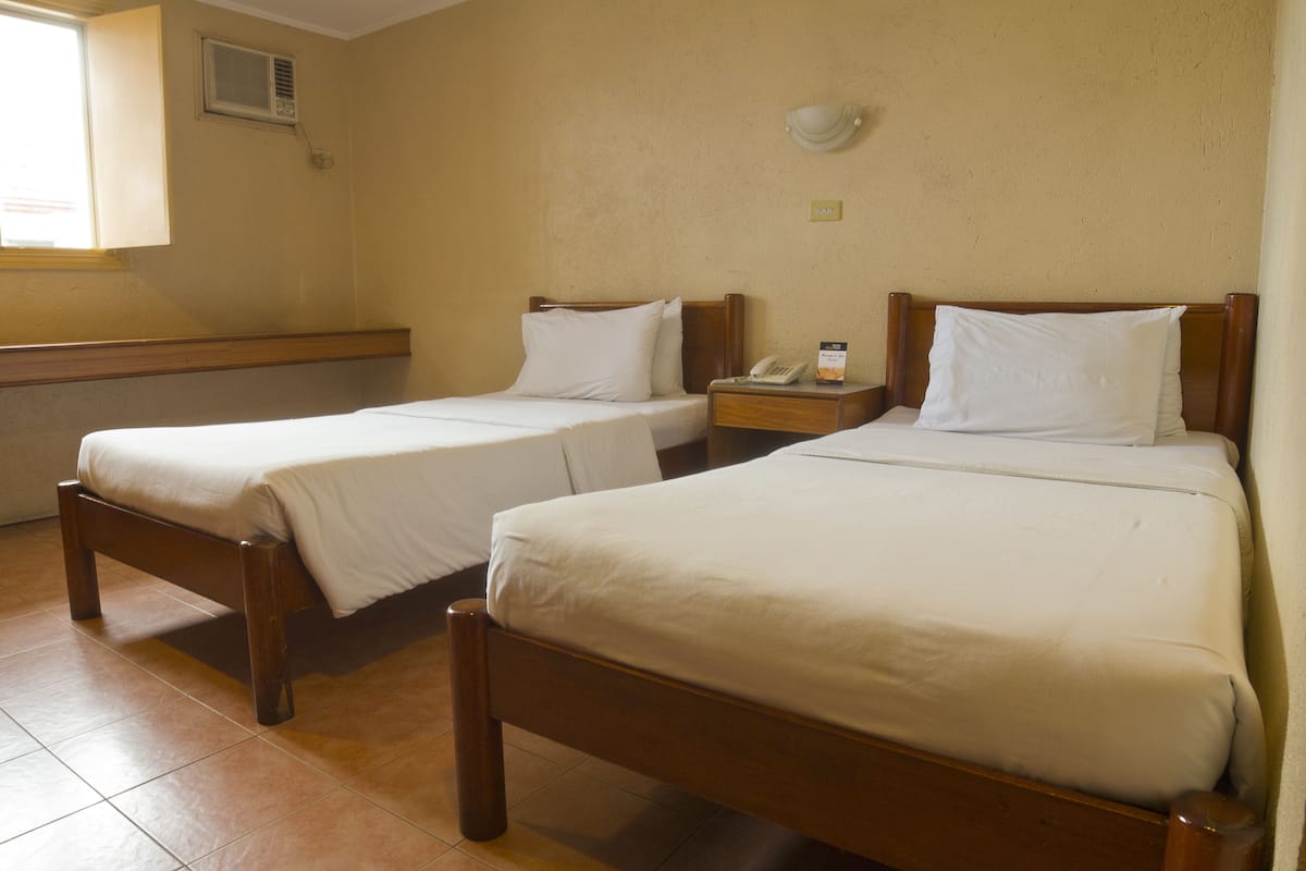Two single beds with wooden frames and white linens in a room with beige walls and a window.