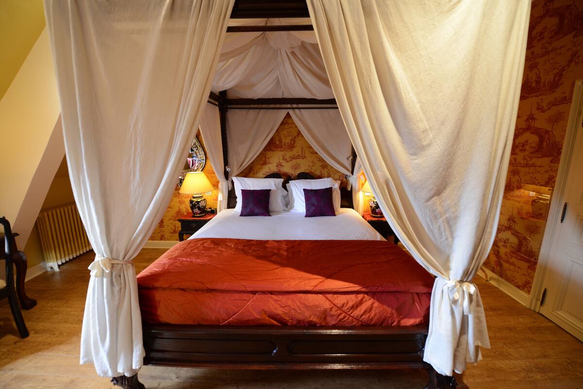 A four-poster canopy bed with a red bedspread, white curtains, and two nightstands with lamps.