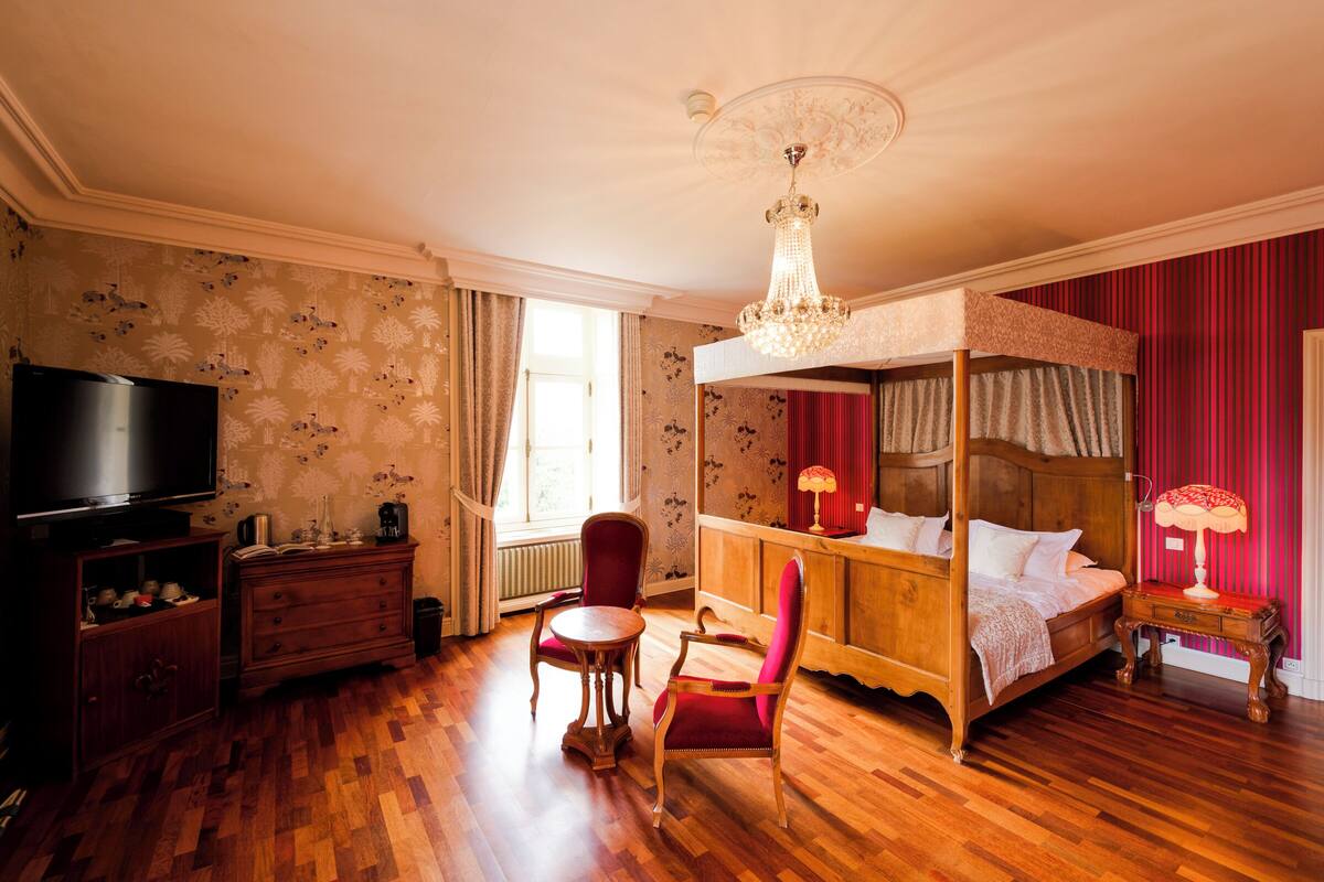 A four-poster bed with a wooden headboard, a television on a wooden cabinet, a red chair, a small round table, and a chandelier.
