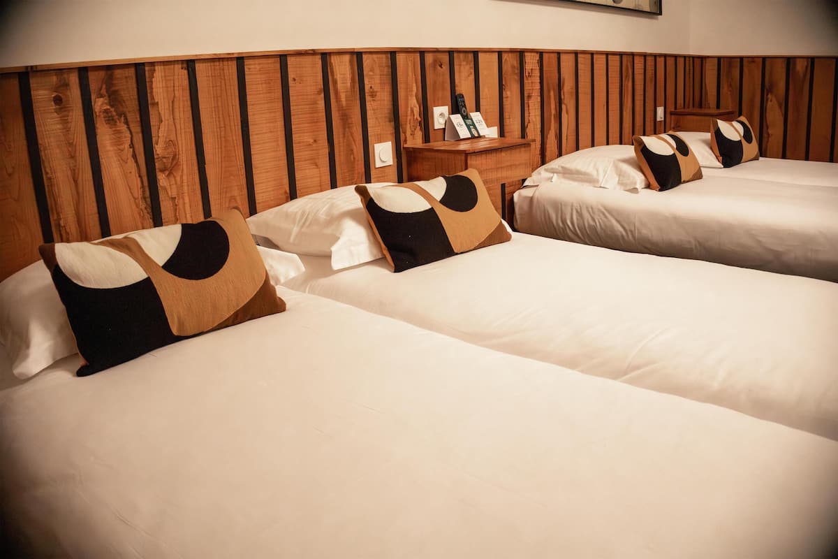 A hotel room with three beds, each with white bedding and decorative pillows. There is a wooden headboard and a small wooden side table with a remote control and a sign.