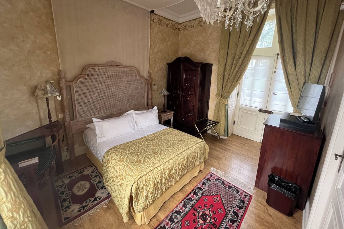 A bedroom with a bed, a wooden wardrobe, a television on a cabinet, a chandelier, and a window with curtains.