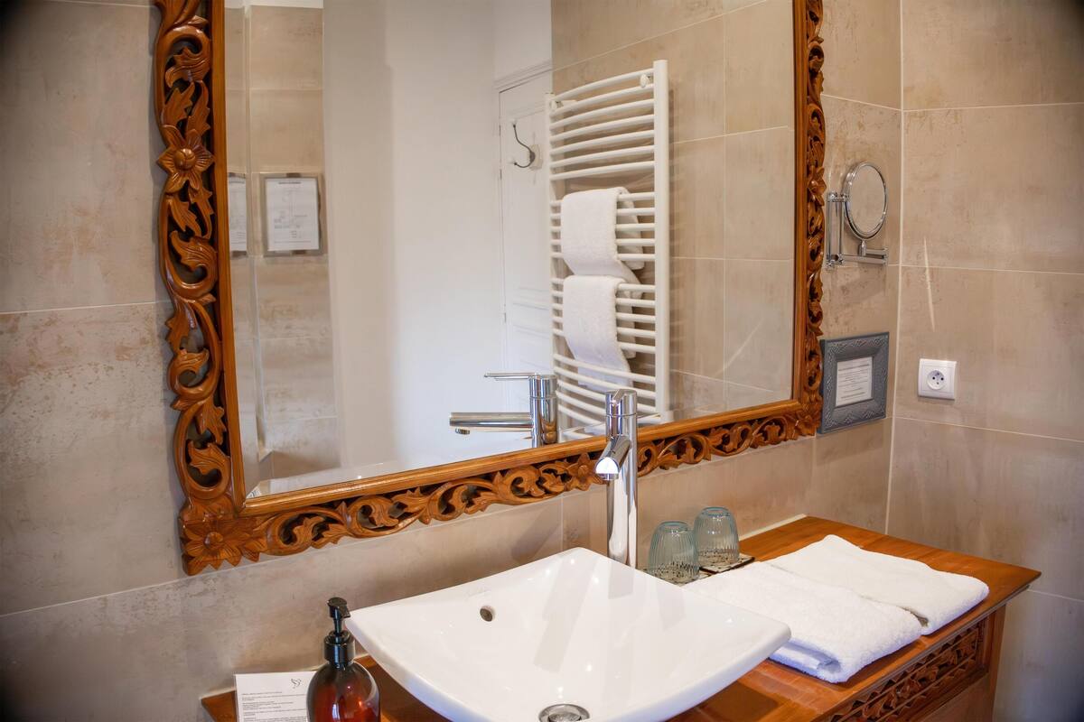 A bathroom with a wooden vanity, a white sink, a large mirror with an ornate frame, a towel rack, and a shower area.