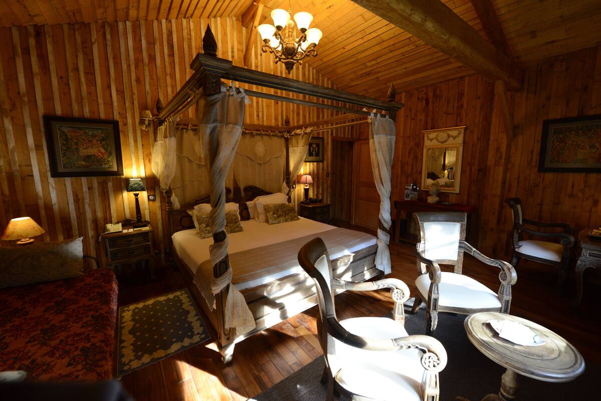 A four-poster canopy bed with a nightstand and lamp, a chair, and a small table in a wooden room.