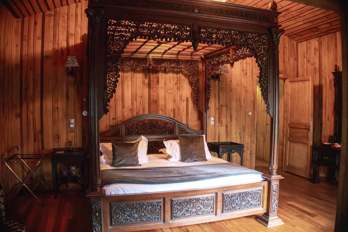 A four-poster bed with intricate carvings, a wooden floor, and a wooden-paneled room.