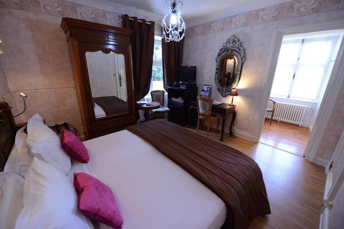 A hotel room with a large bed, a television, a desk with a chair, a mirror, and a chandelier.