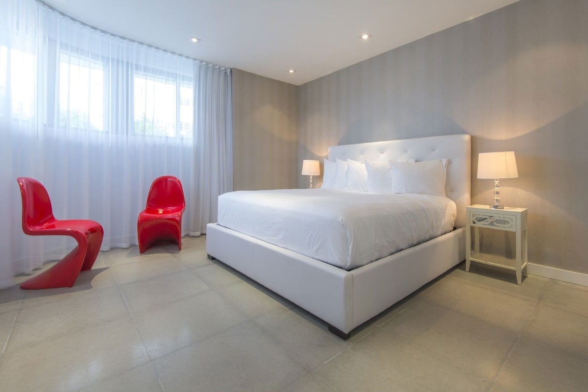 A modern hotel room with a large bed, two red chairs, a nightstand with a lamp, and a window with sheer curtains.