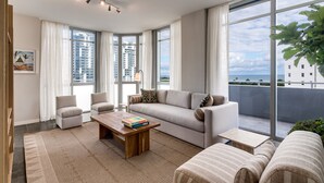 Ein modernes Wohnzimmer mit einer Couch, Sesseln, einem Couchtisch und großen Fenstern mit Blick auf die Stadt und den Ozean.