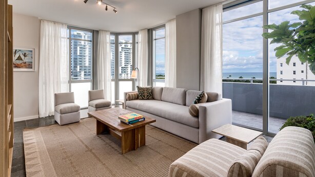 Uma sala de estar moderna com sofá, poltronas, mesa de centro e grandes janelas com vista para a cidade e o oceano.
