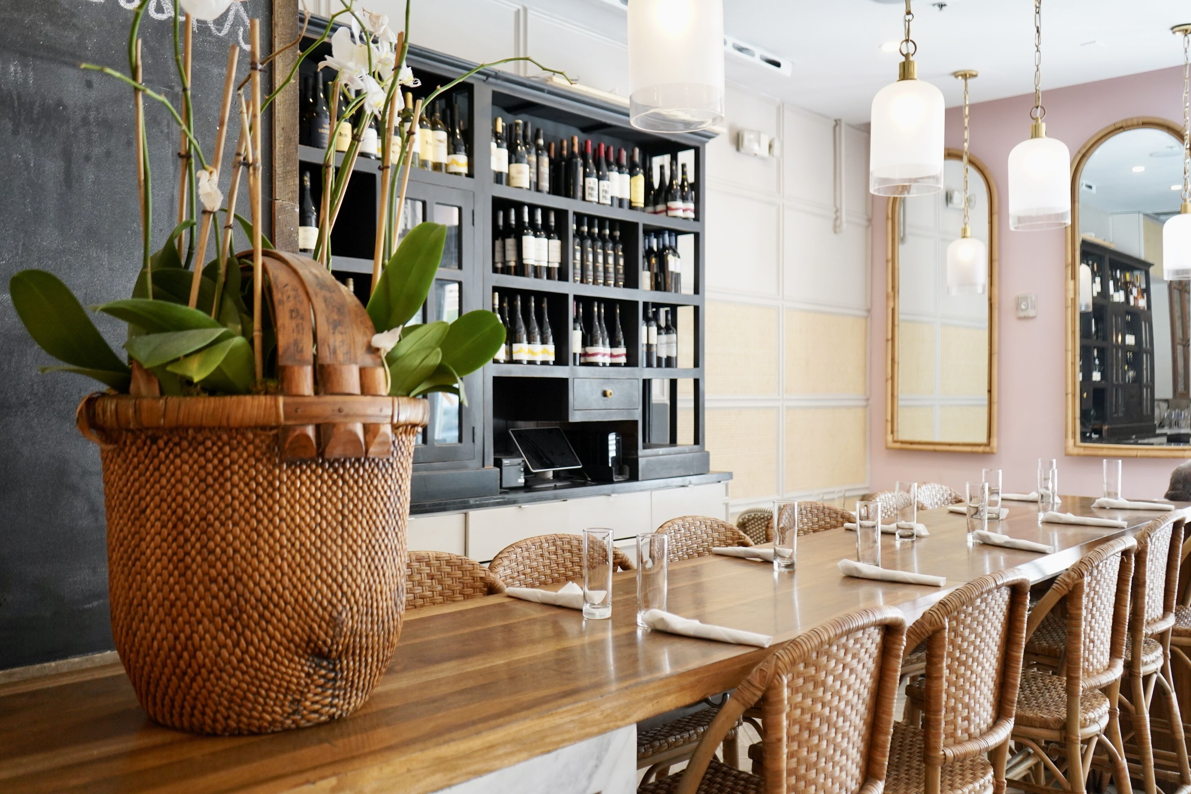 A dining area with a long wooden table, wicker chairs, and a large woven plant centerpiece.