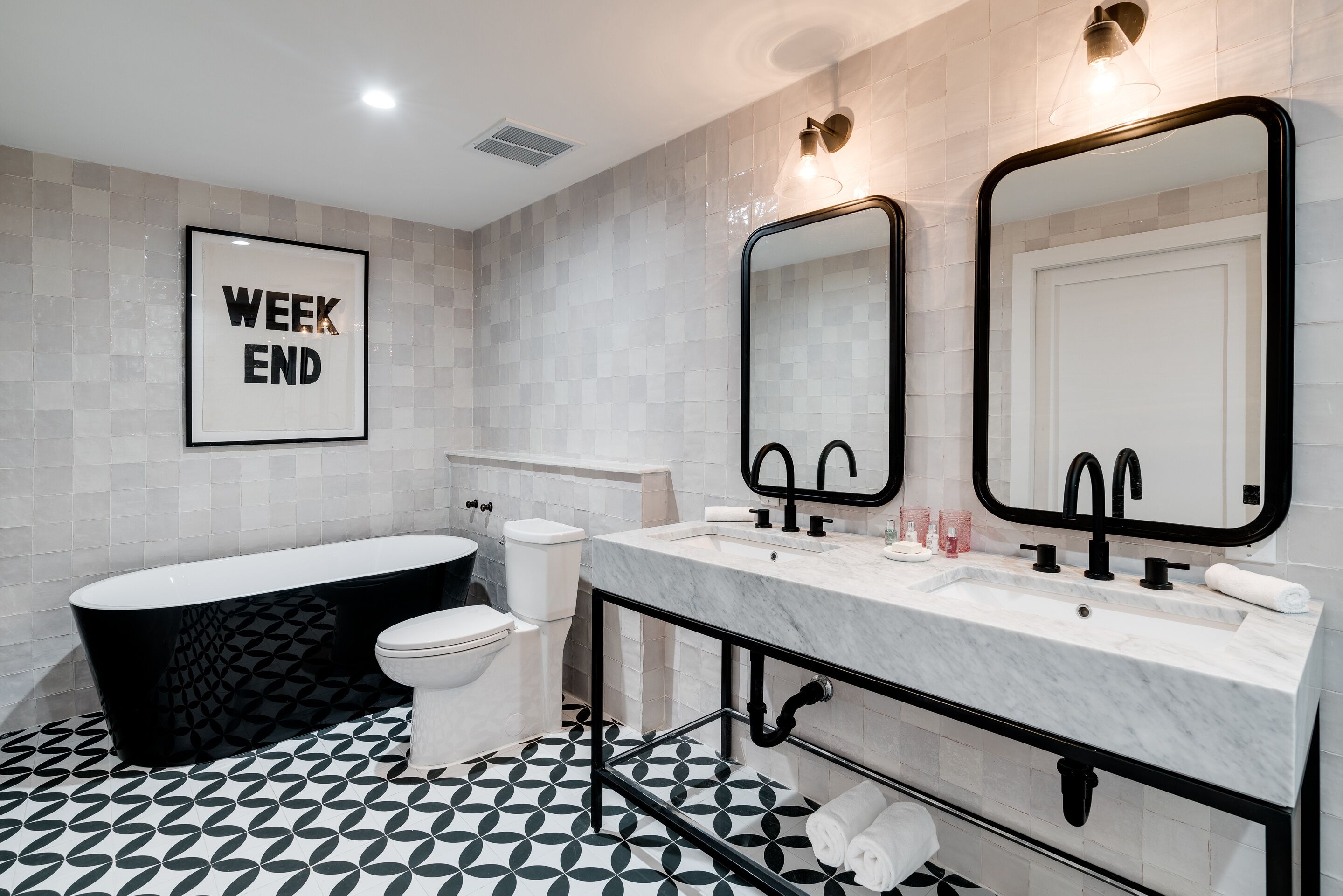 Un baño moderno con suelo de baldosas blanco y negro, una bañera independiente, un inodoro, un tocador con dos lavabos y un espejo grande.