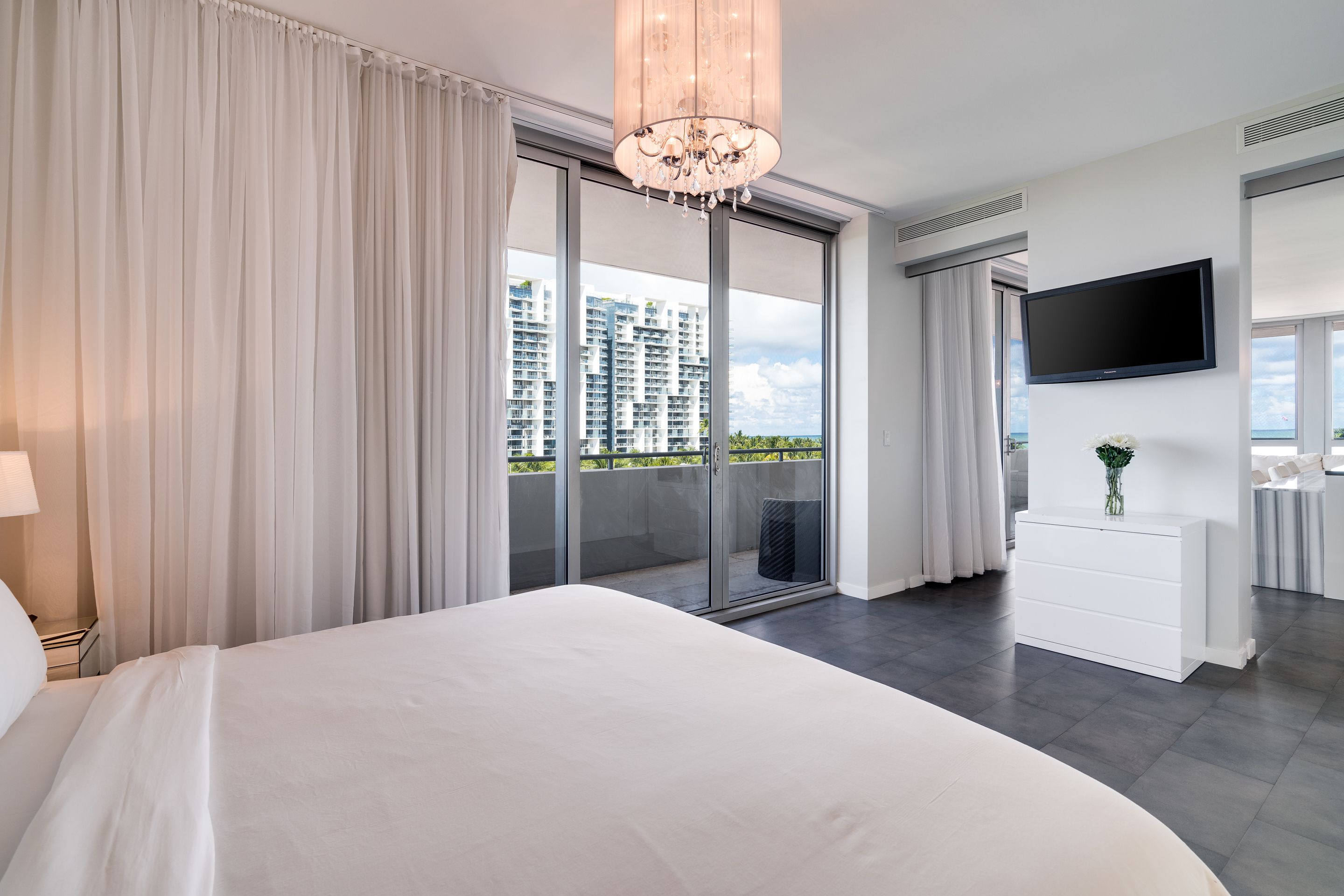 Habitación de hotel moderna con una cama grande, una araña, un televisor de pantalla plana, un aparador blanco y un balcón con vista a la ciudad.