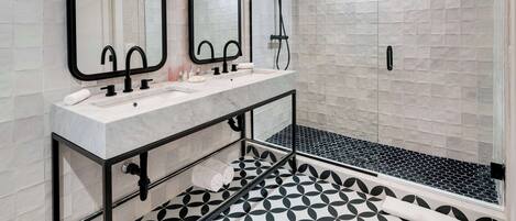 A modern bathroom with a large mirror, two sinks, a shower, and a patterned floor.