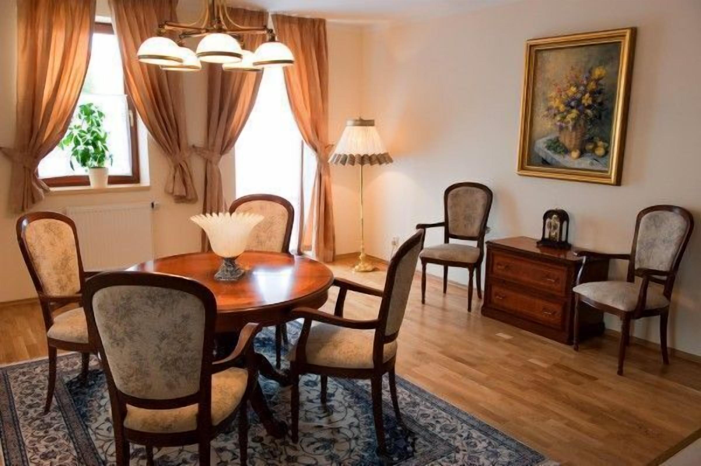 A dining room with a round wooden table, six upholstered chairs, a chandelier, a floor lamp, a painting on the wall, and a wooden cabinet.