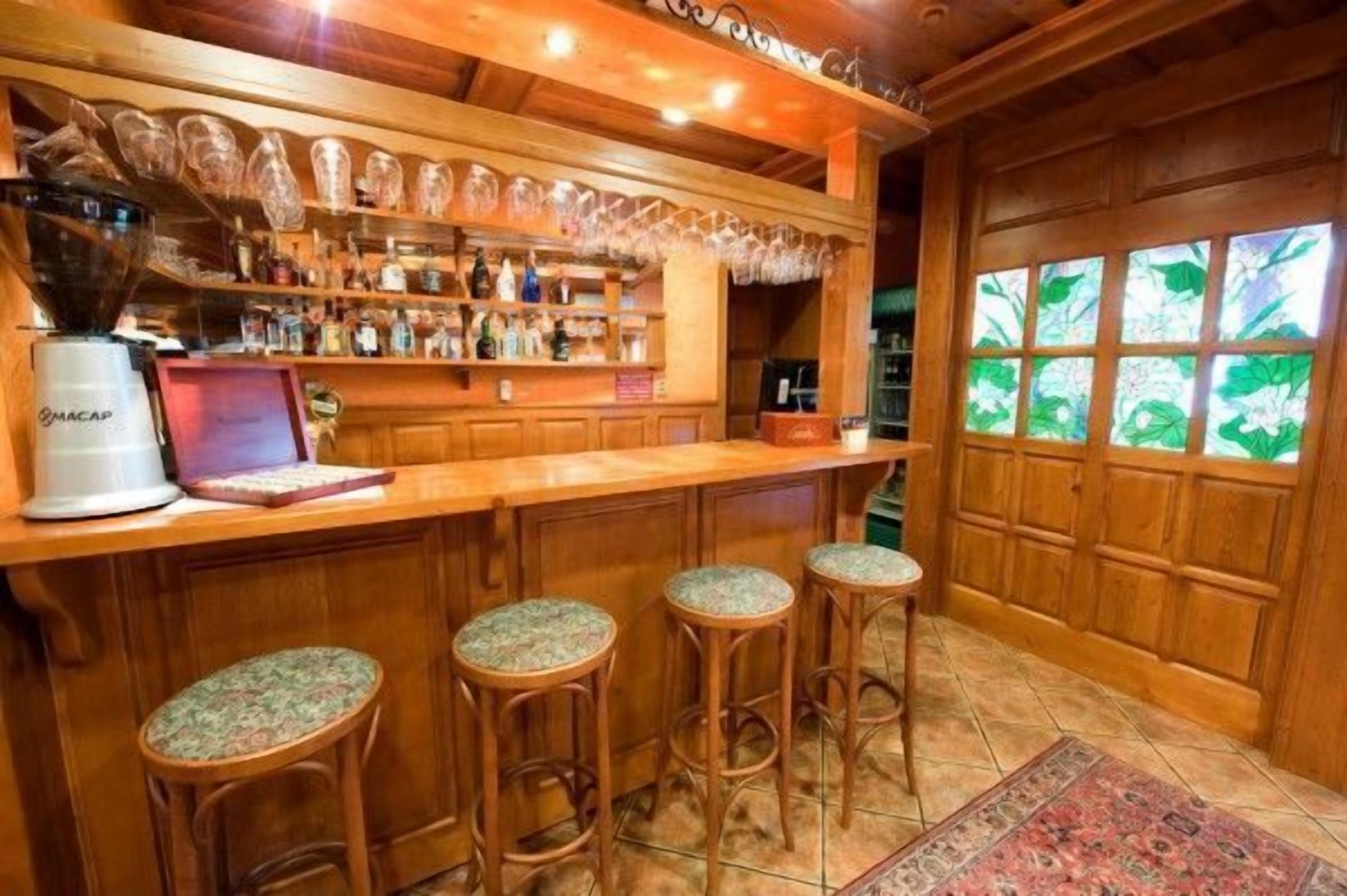 Uma área de bar com balcão de madeira, banquetas, uma máquina de café e prateleiras com garrafas e copos.