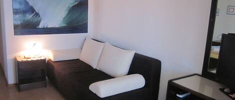 A room with a dark-colored sofa, a small bedside table with a lamp, a large framed picture of a wave, a small framed picture on the wall, and a mirror on a dresser.