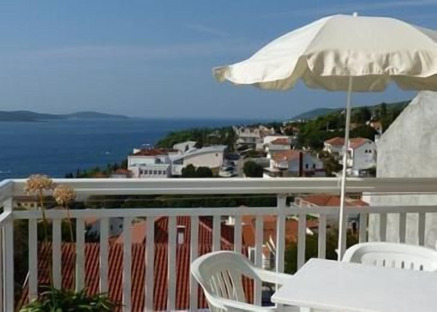A balcony with a white umbrella, white chairs, and a view of the sea and houses.